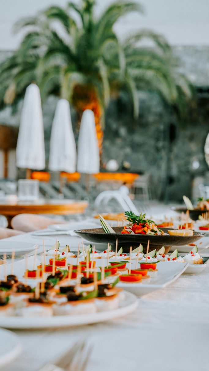 A variety of elegantly arranged appetizers on a table outdoors with palm trees in the background.