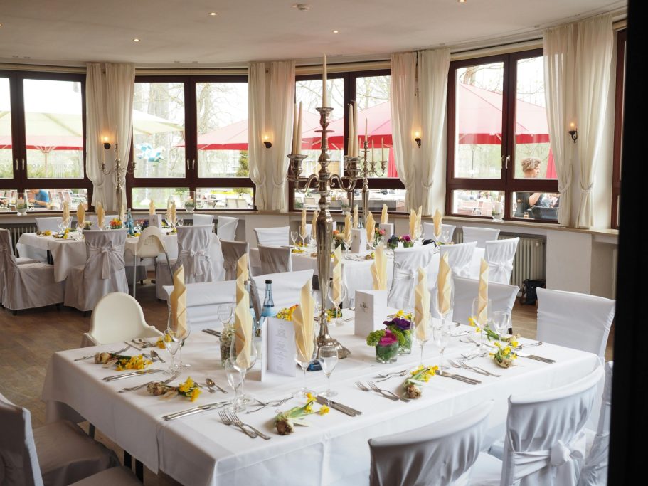 Elegant dining setup with white tablecloths, flowers, and large windows in a bright room.
